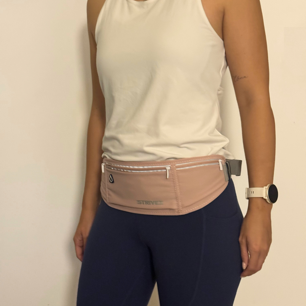 A person wearing a light pink Strive running waist bag over a white tank top and navy blue leggings, standing against a plain off-white wall.